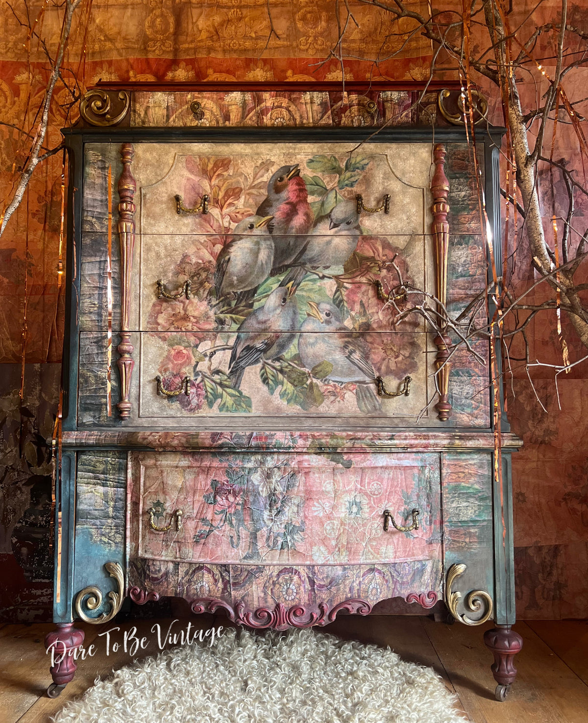 A distressed vintage-style wooden cabinet or hutch with a large painted panel showing birds and flowers, set against a rustic wood wall with decorative branches and a white shag rug.